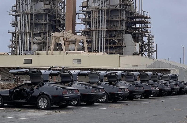 lineup of matte black DeLorean cars with gull-wing doors open, parked in front of an industrial power plant structure with pipes and metal frameworks in the background